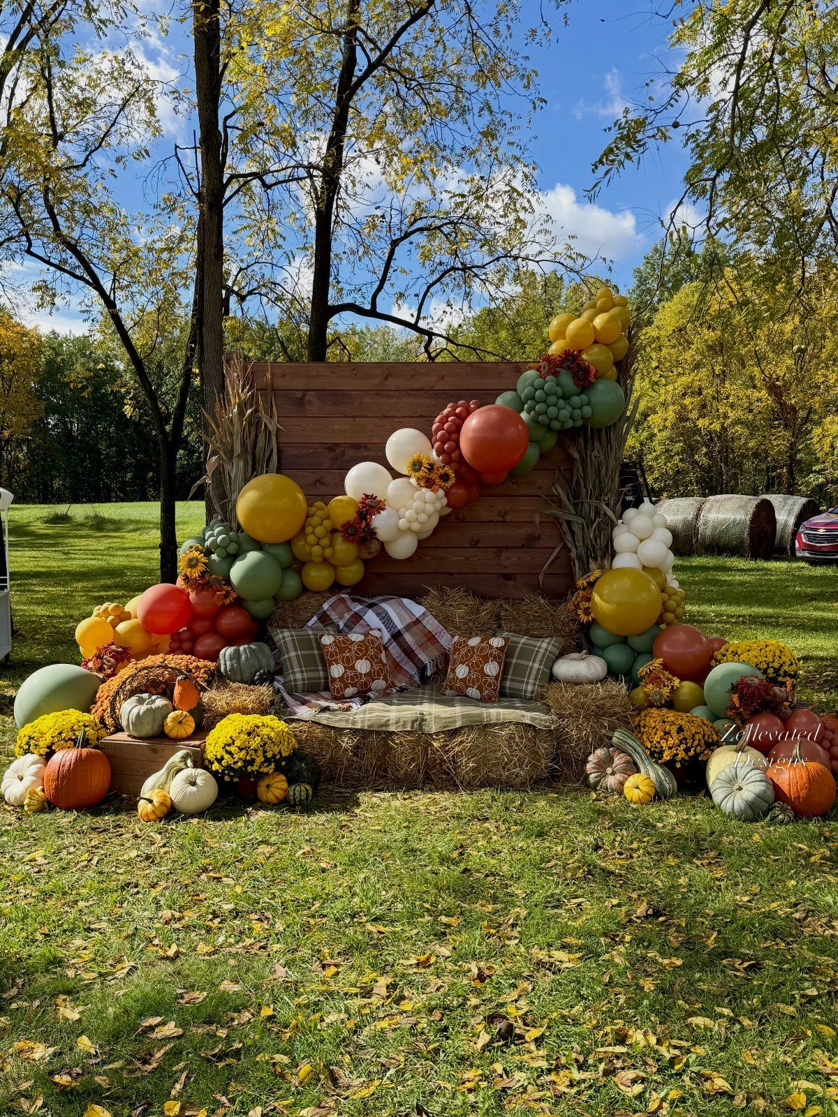Autumn photo backdrop with hay bales, pumpkins, and a colorful balloon arch outdoors.