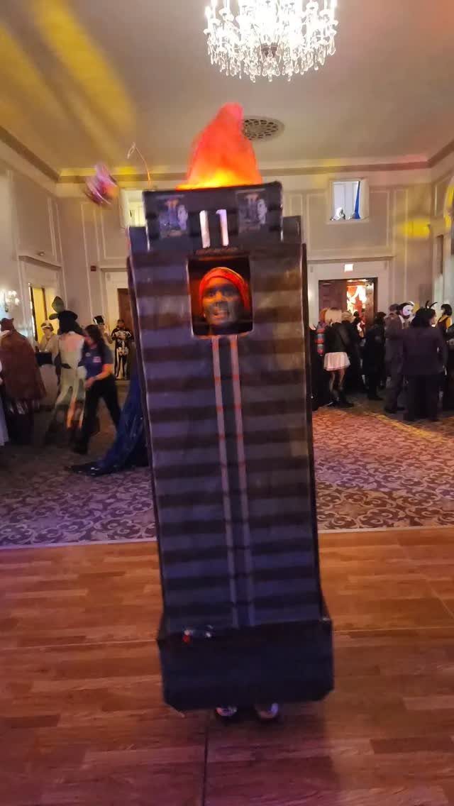 Person in a skyscraper costume with a flame on top at a party.