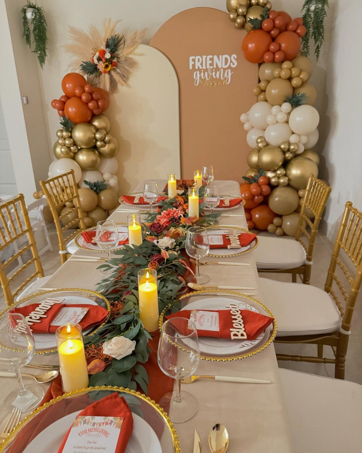 Friendsgiving table setting with gold chairs, red napkins, candles, and balloon arch backdrop.