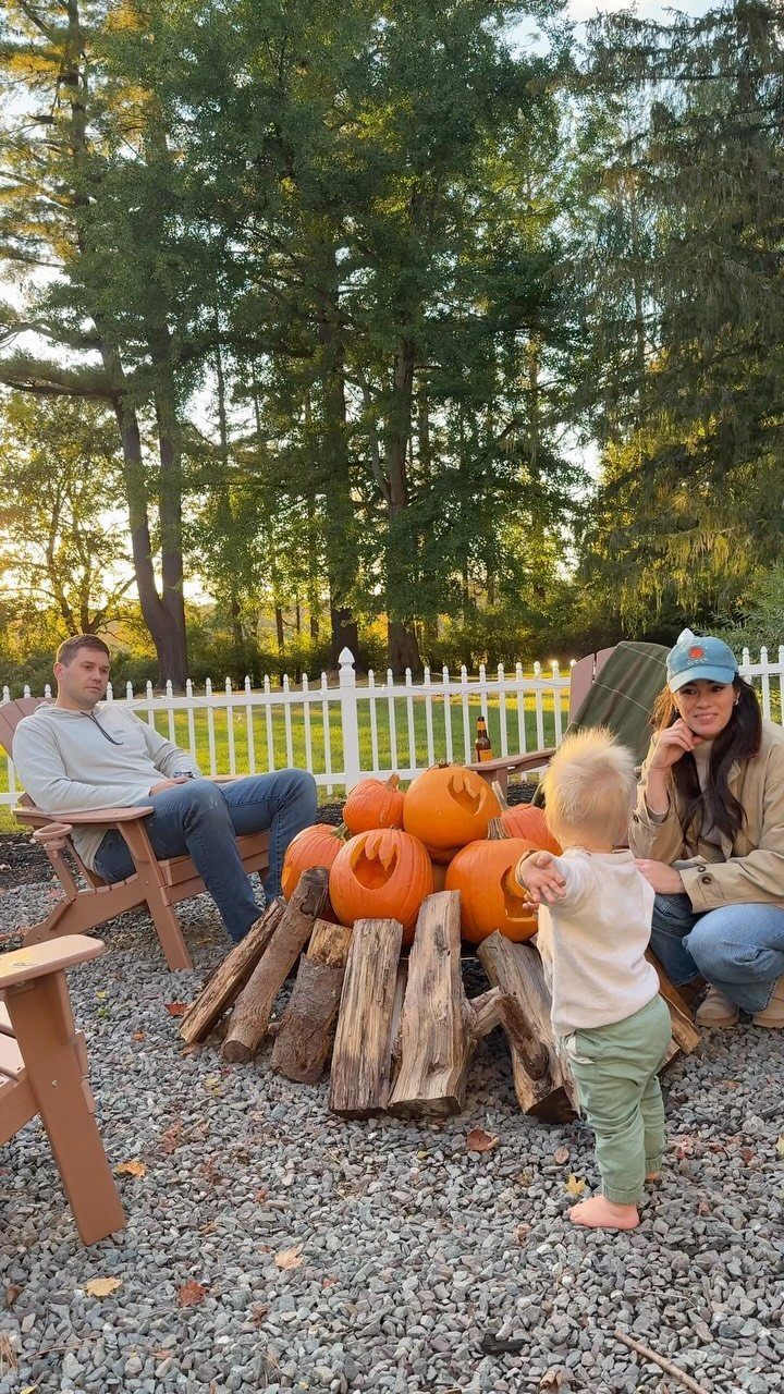 Family gathering around a log fire pit filled with orange pumpkins in a backyard.