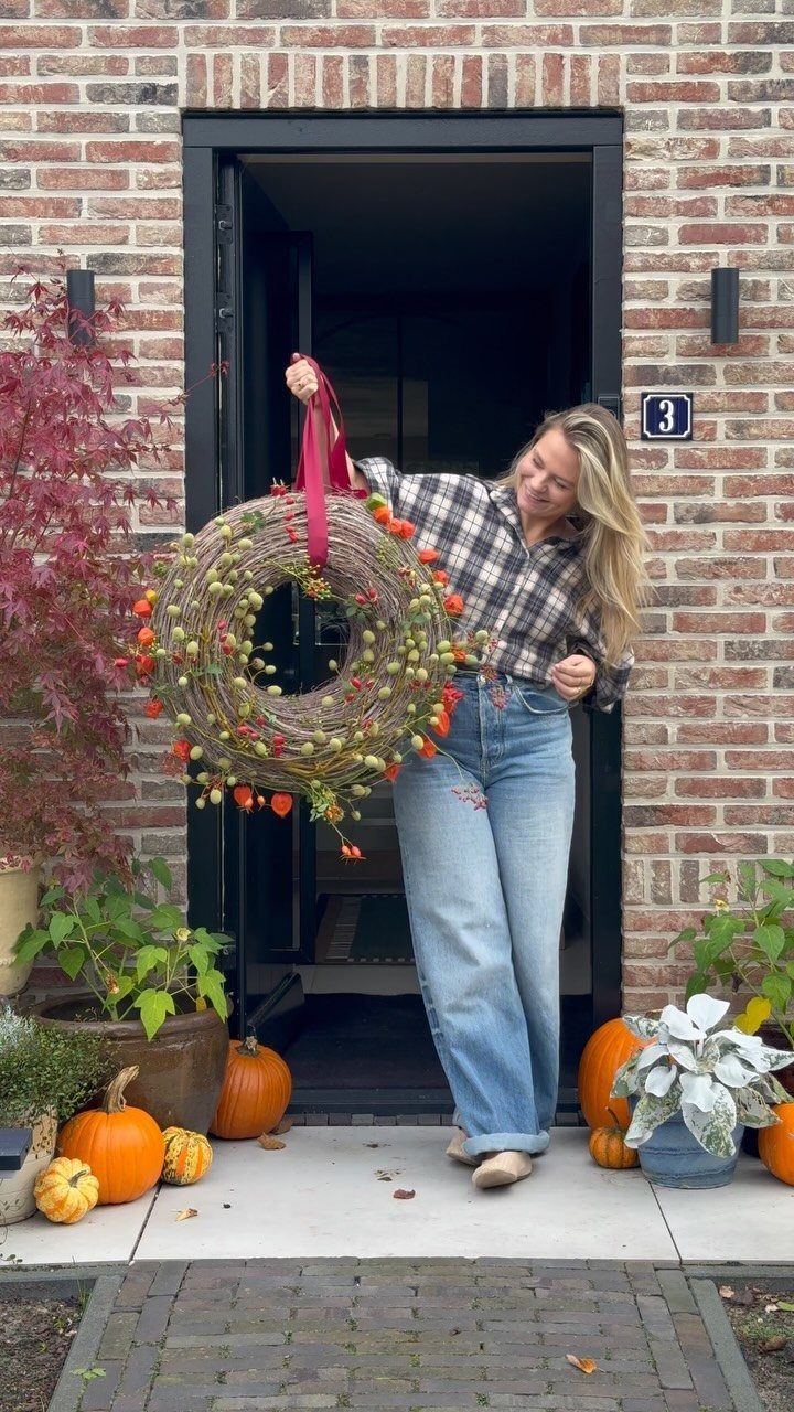 Woman holding a handmade autumn twig wreath in a brick house doorway with pumpkins.