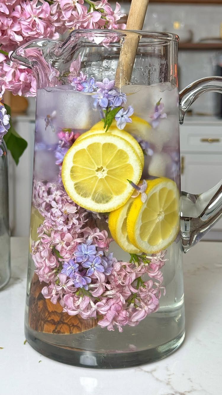 Glass pitcher of water infused with lemon slices and pink and purple lilac flowers.