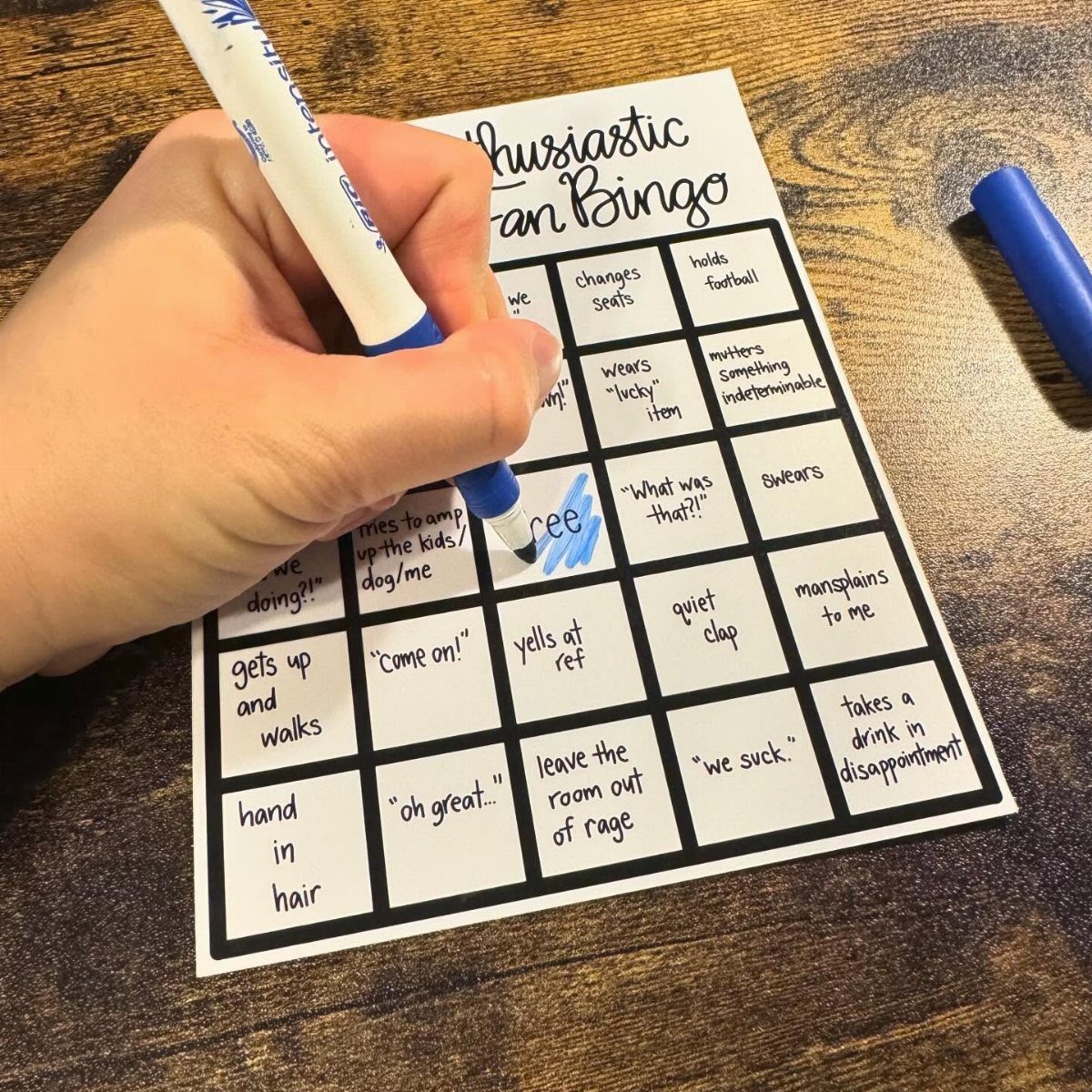 Hand marking a fan bingo card on a wooden table with blue marker