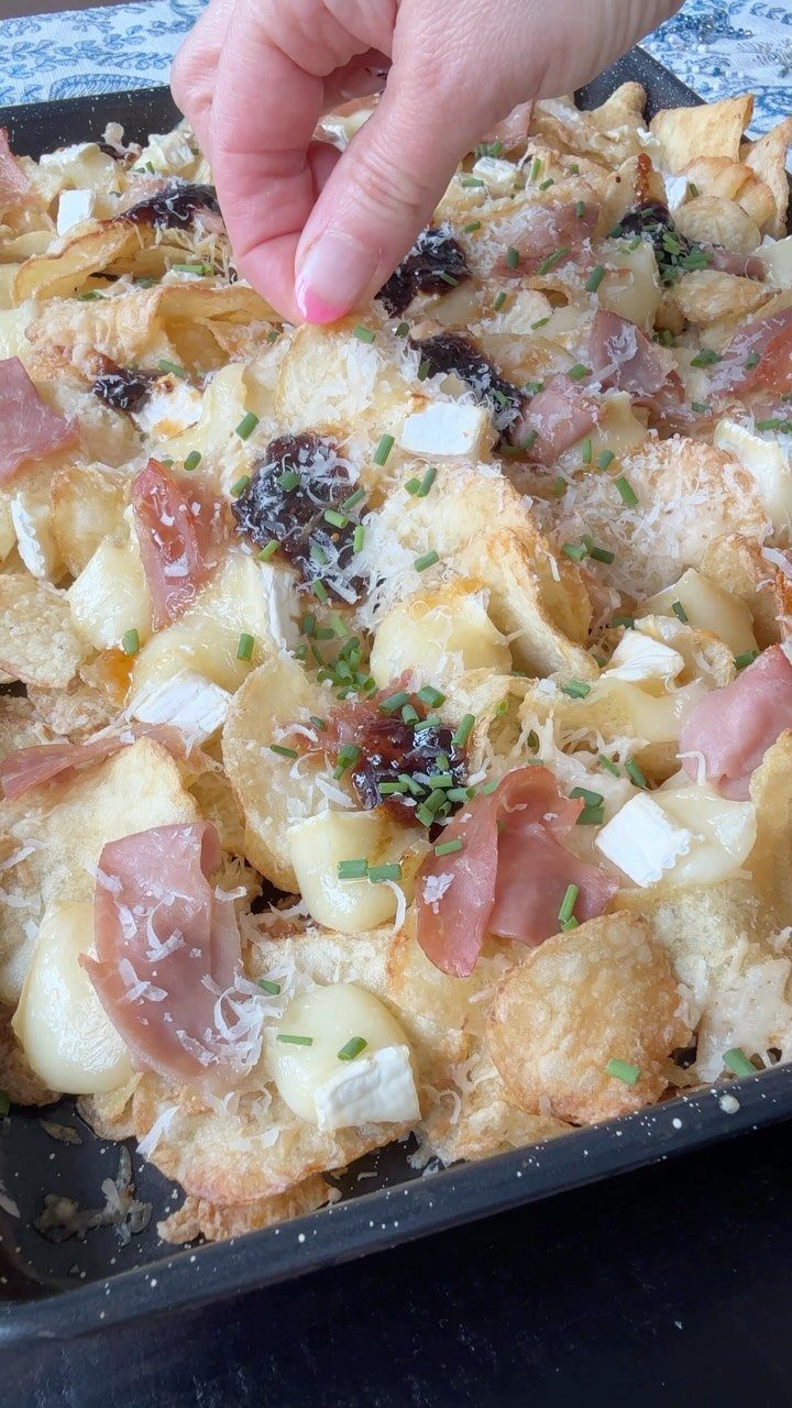 Potato chips with cheese, prosciutto, and chives on a black baking sheet.