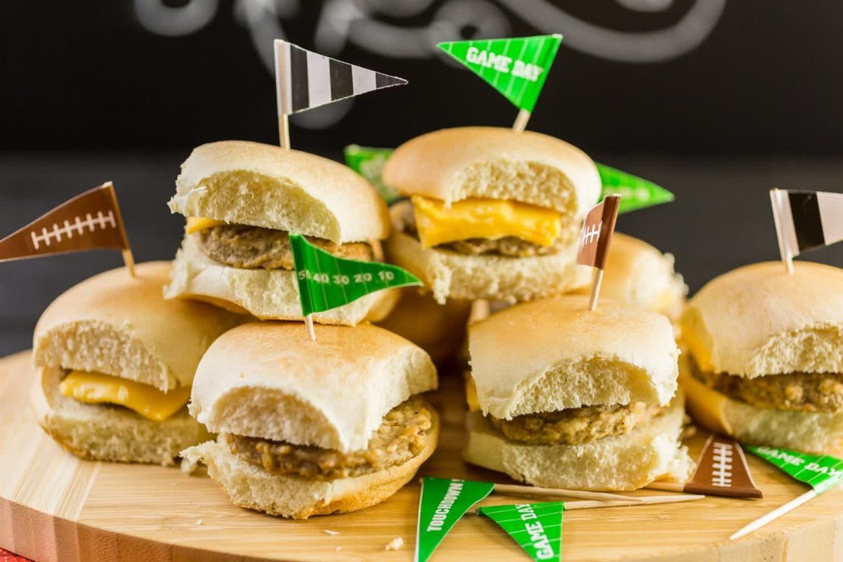 Mini sliders with cheese and meat patties decorated with football-themed flags on wood.