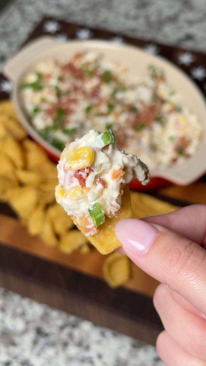Creamy corn and bacon dip on a corn chip held by a hand.