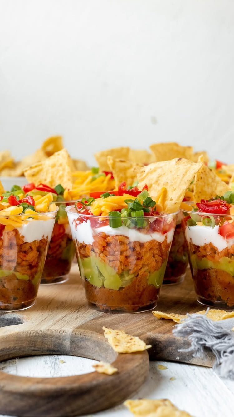 Layered bean and guacamole dips in clear cups with tortilla chips on wood.