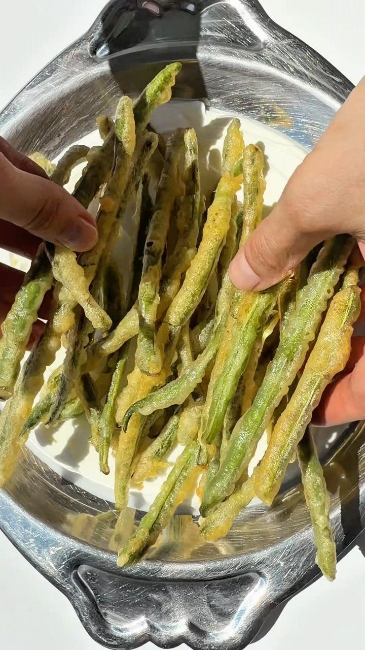Fried green beans in a silver platter being picked up by two hands.