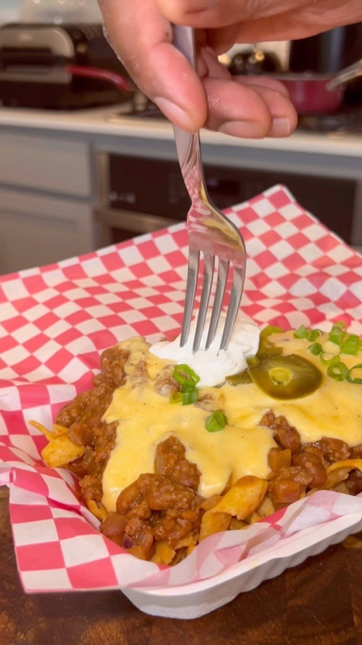 Frito chili pie with melted cheese and jalapeños in a checkered paper tray.