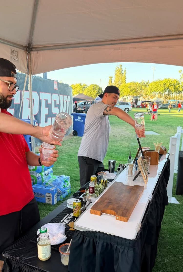 Men preparing drinks at a catered table under a tent at a tailgate party.