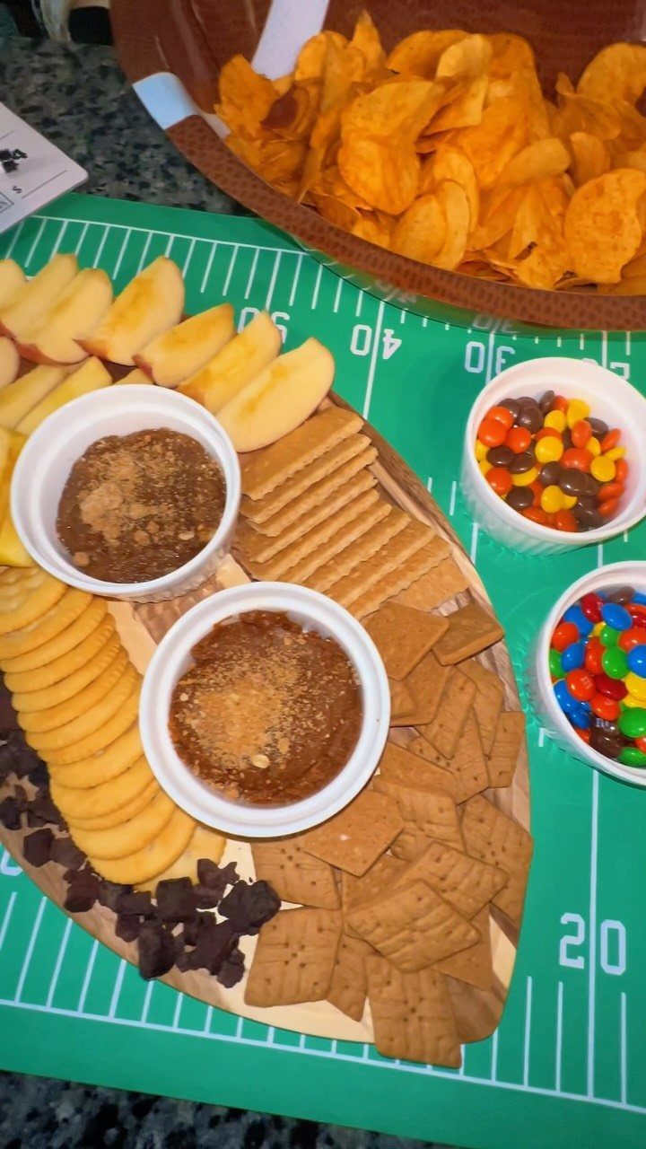 Football shaped dessert board with apples, crackers, and dips on a field tablecloth.