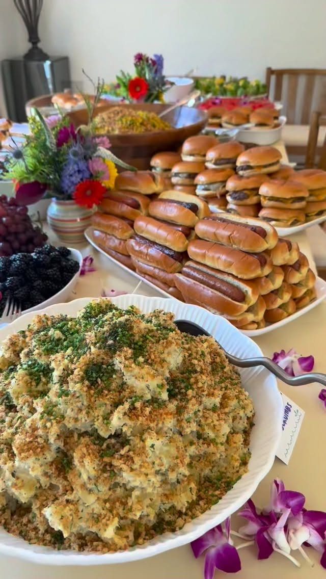 Platters of sliders and hot dogs alongside potato salad and fresh fruit.