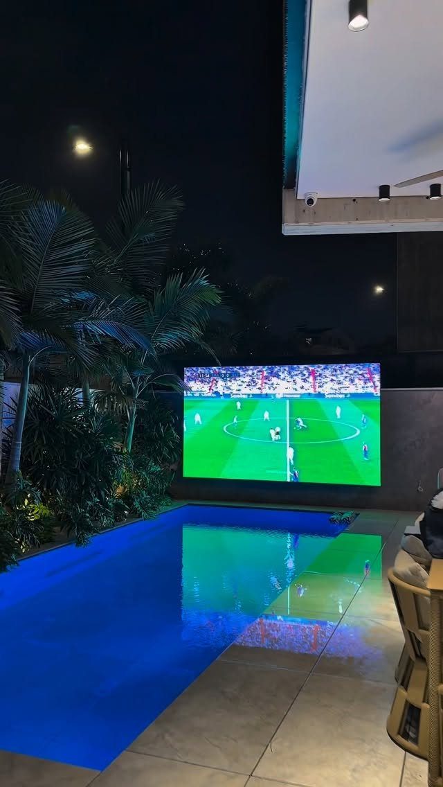 Outdoor screen showing a football match next to a glowing blue swimming pool