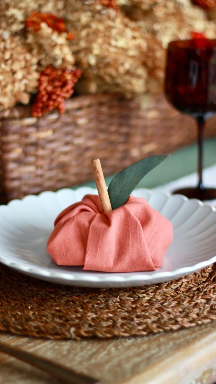 Orange cloth napkin folded like a pumpkin on a white scalloped plate.