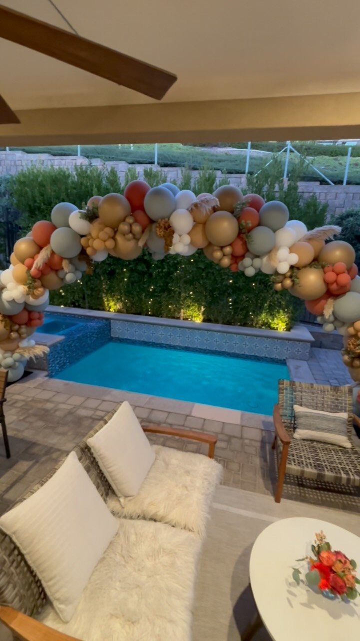 Balloon arch in gold and red colors over a swimming pool patio area.