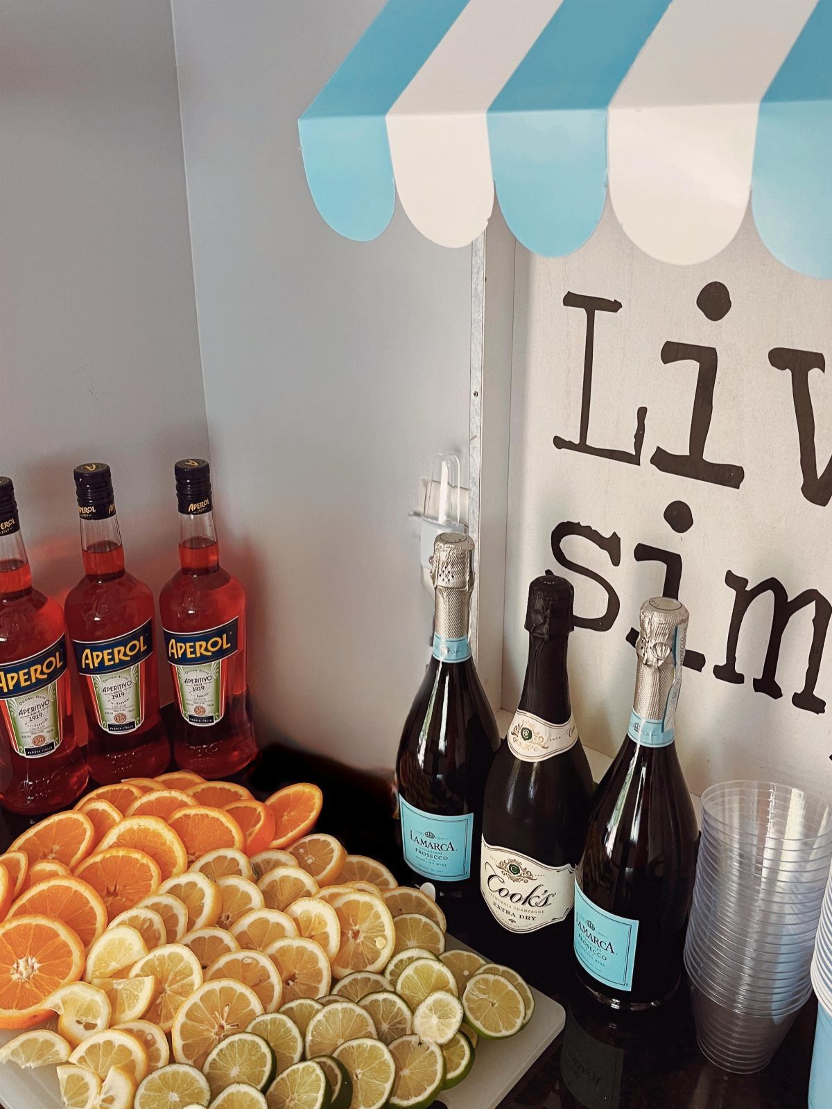 Drink station with Aperol, sparkling wine, citrus slices, and clear plastic cups.