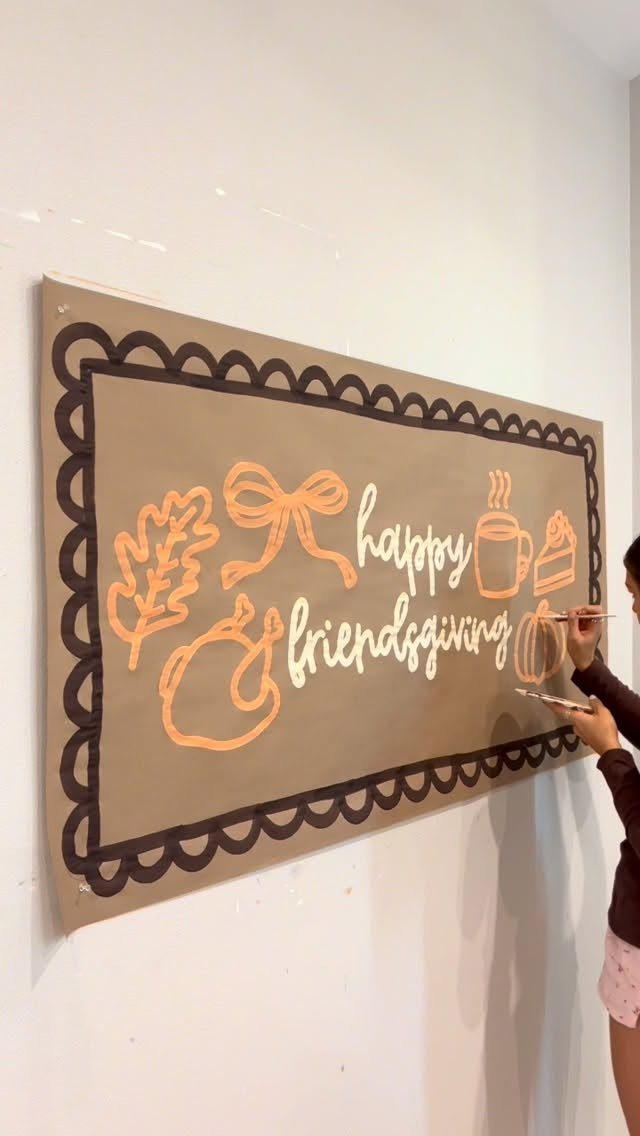 Person painting a tan Friendsgiving banner with orange autumn illustrations on a wall.
