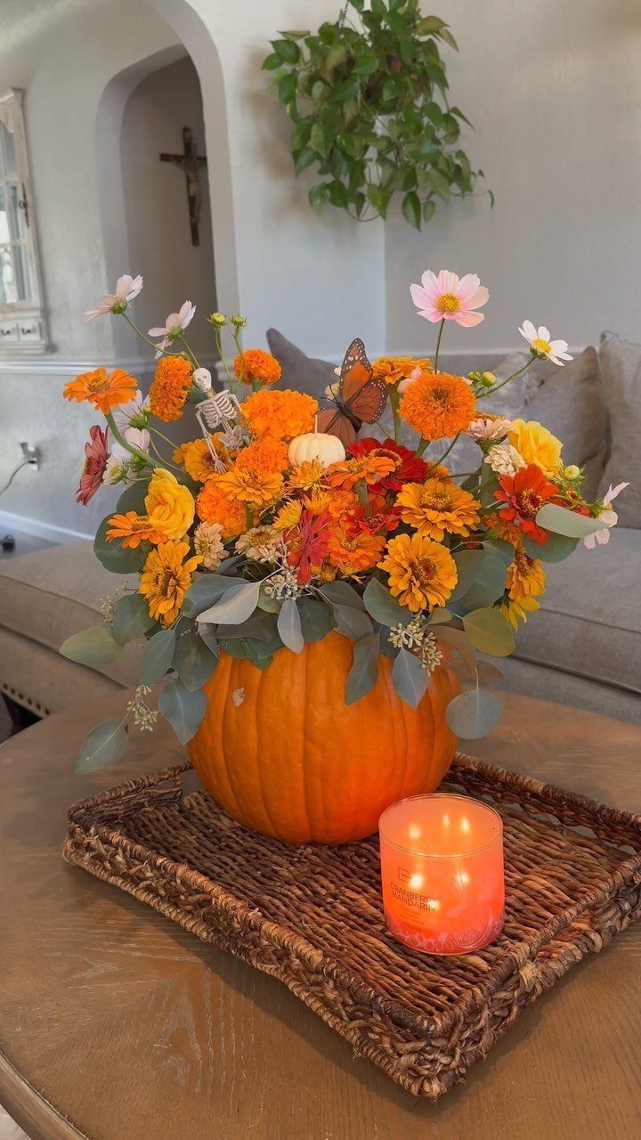 Autumn flowers and greenery arranged inside a pumpkin on a woven tray.