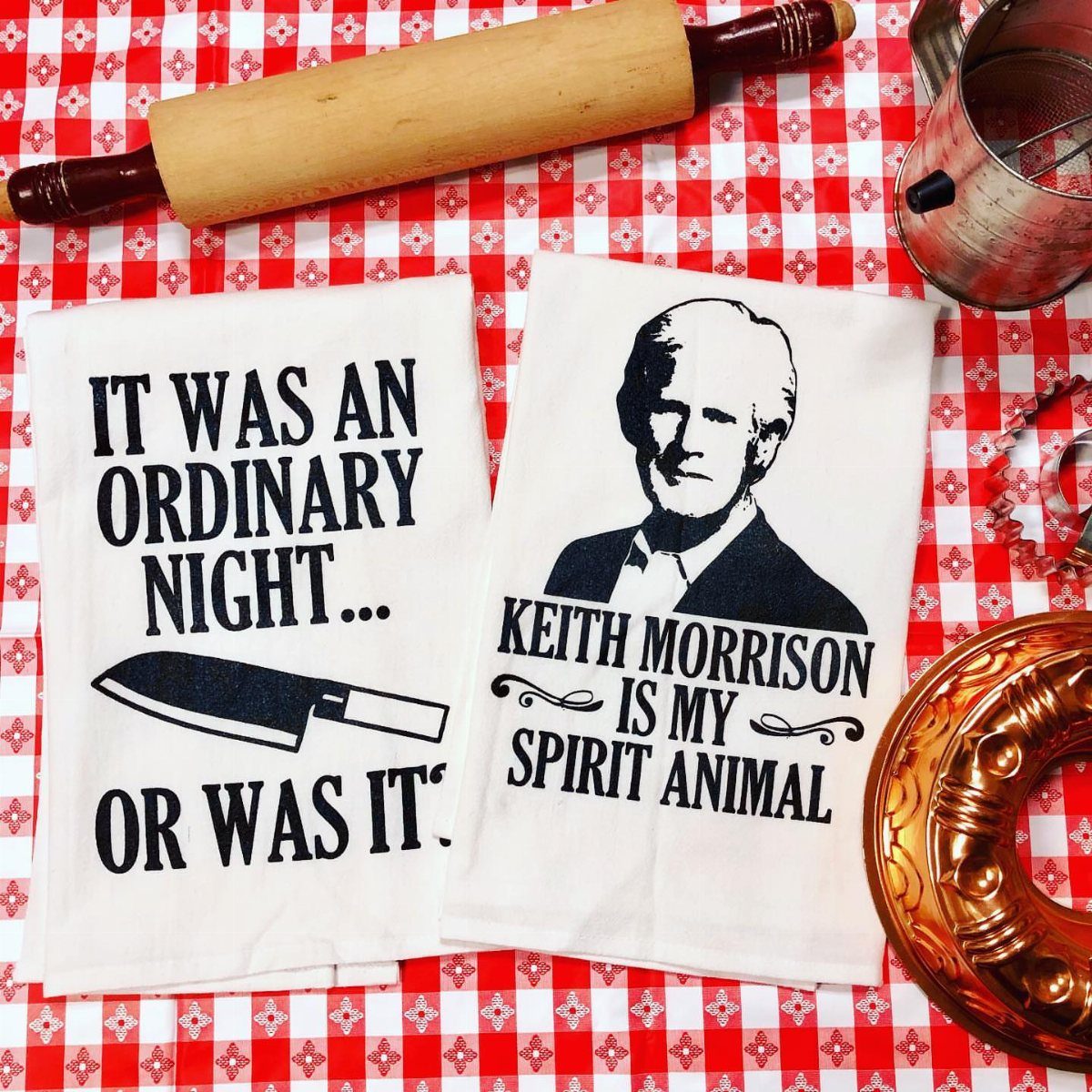 Two white humorous kitchen towels on a red checkered tablecloth with a rolling pin.