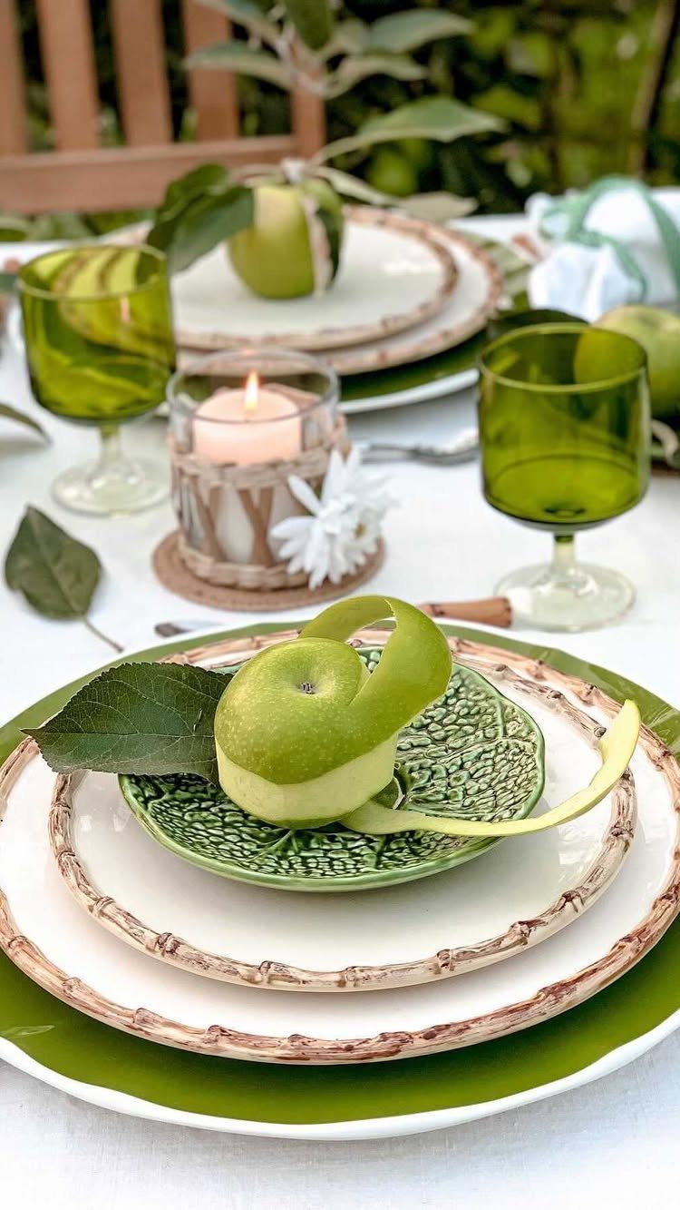Rustic table setting with green apples on white plates and green glassware outdoors.