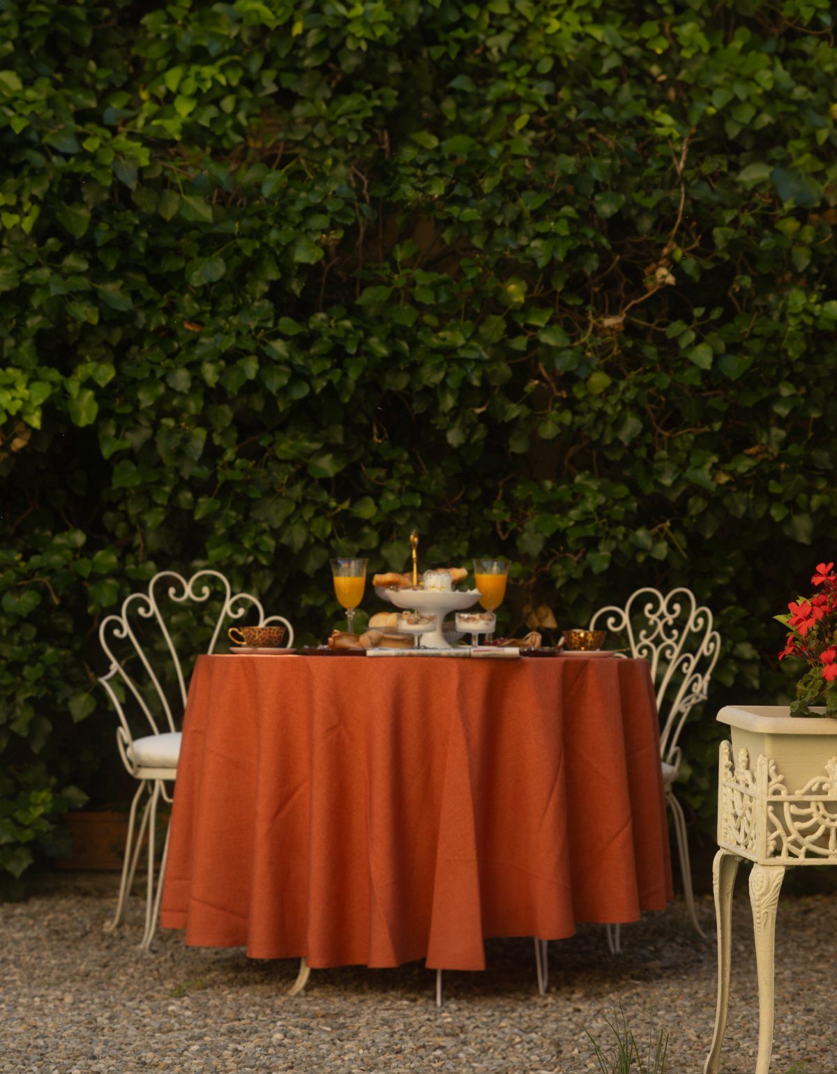 Round table with orange tablecloth and white ornate chairs against a green hedge.