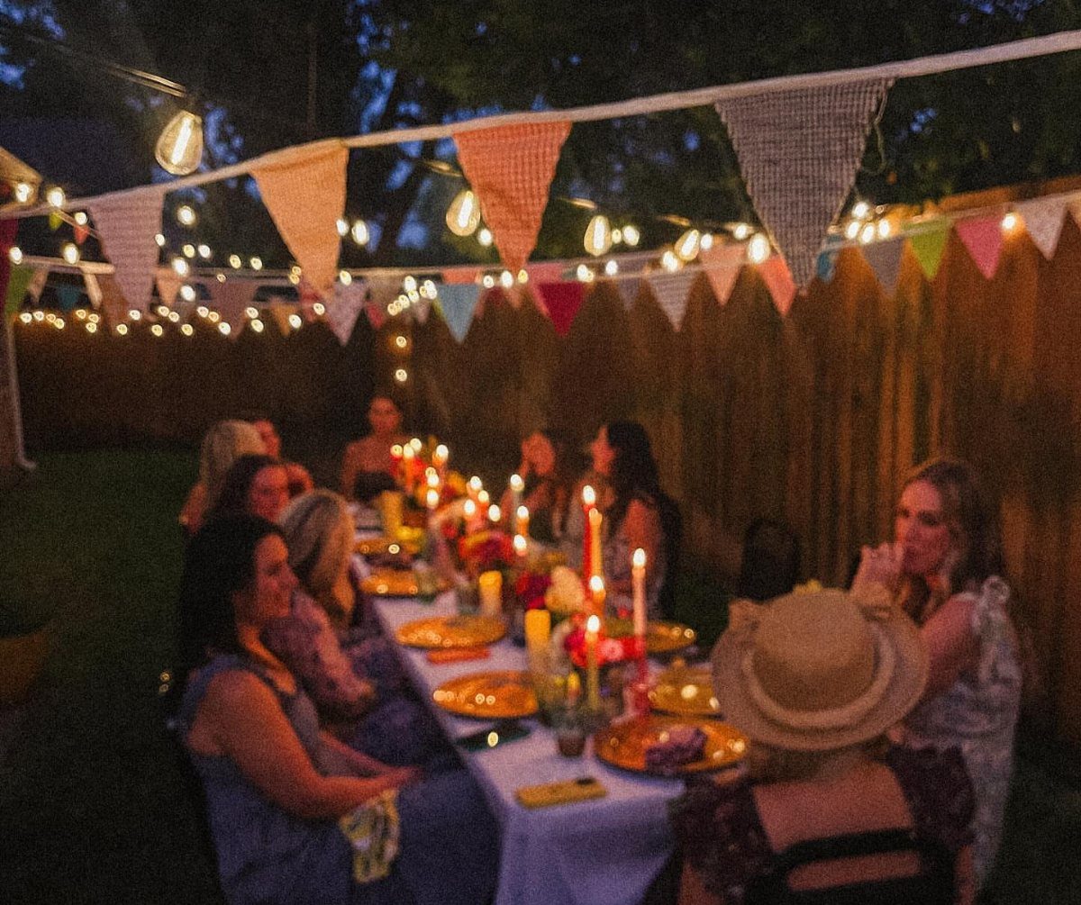 People dining at a long outdoor table with string lights and colorful bunting banners.