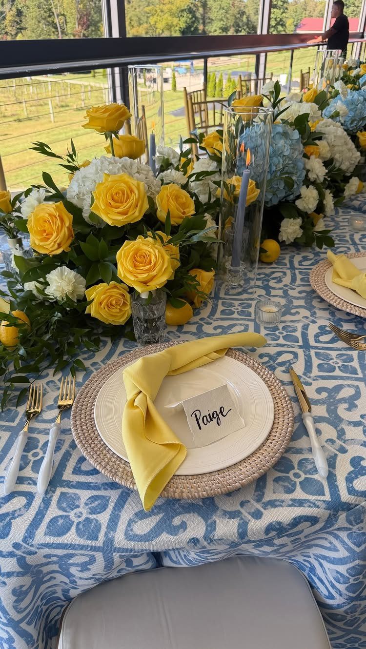 Table setting with yellow roses, blue hydrangeas, and a blue patterned tablecloth outdoors.