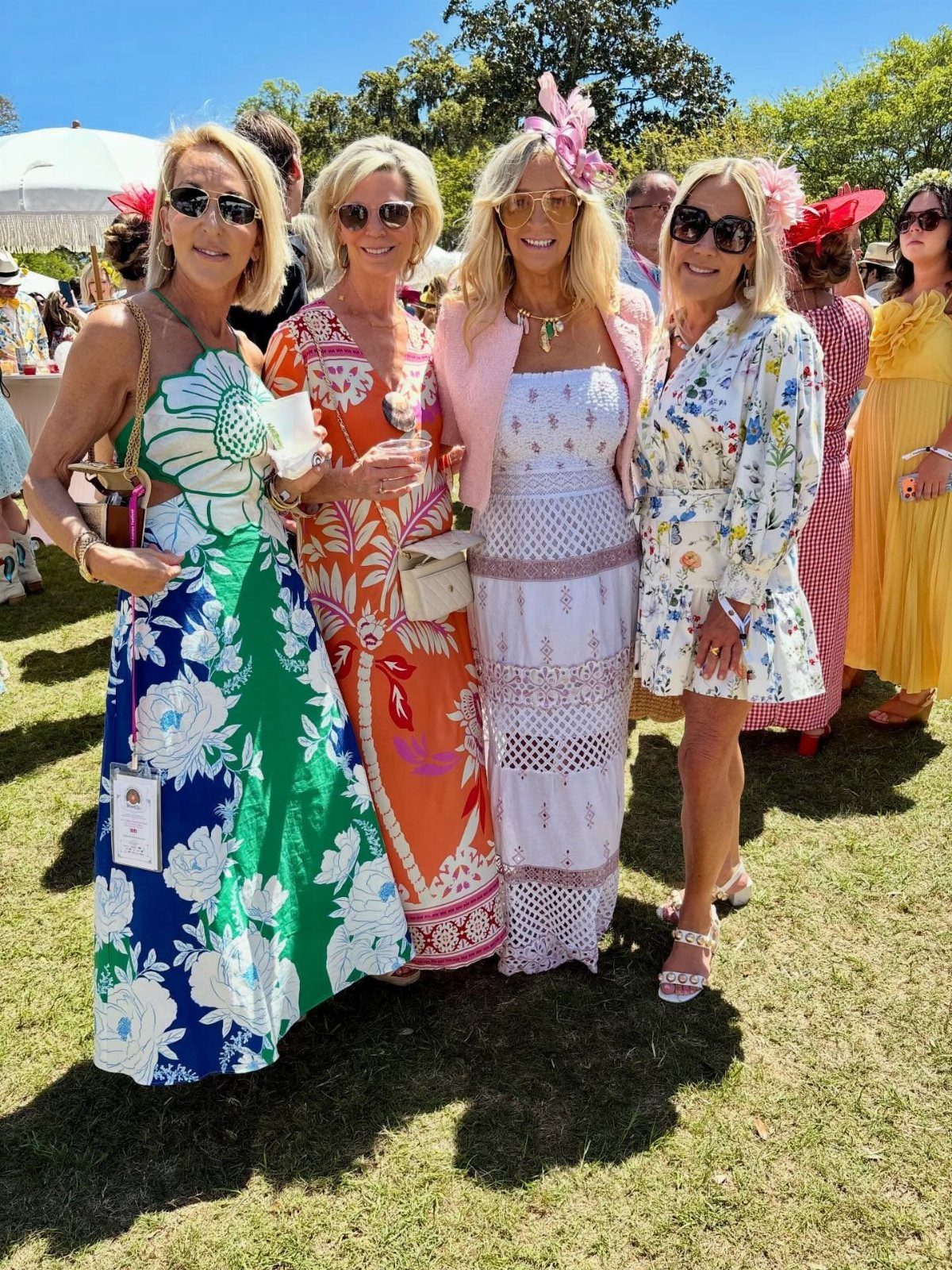 Four women in colorful floral summer dresses posing on a sunny grassy lawn.