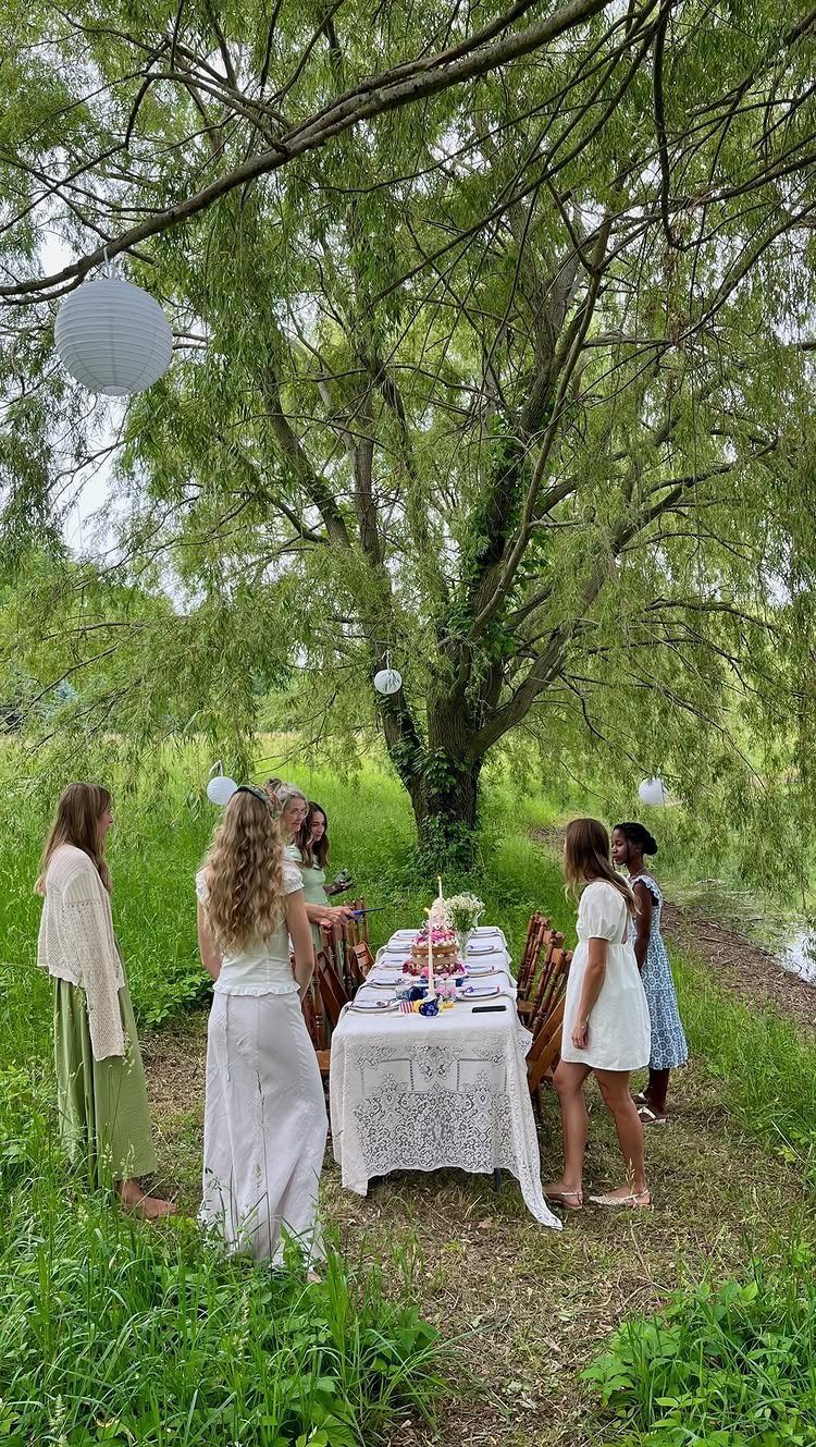 Women gathered around a lace-covered table under a weeping willow with paper lanterns.