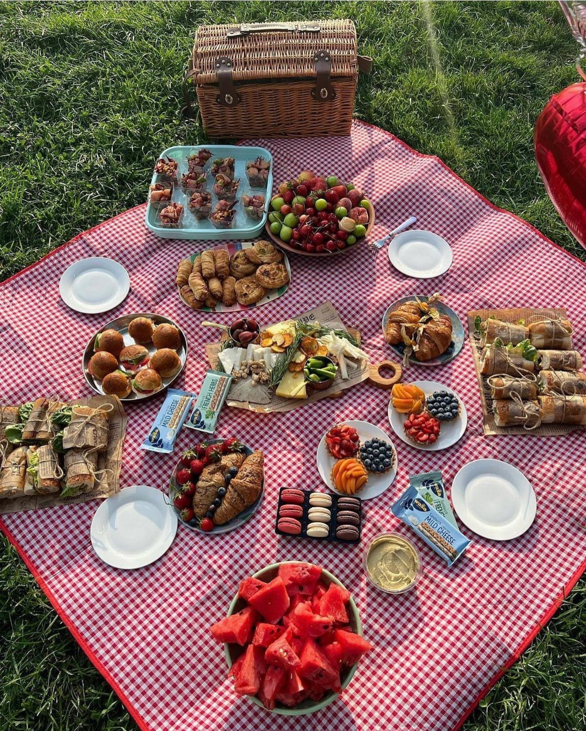 Picnic blanket on grass with a wicker basket and various gourmet snacks.