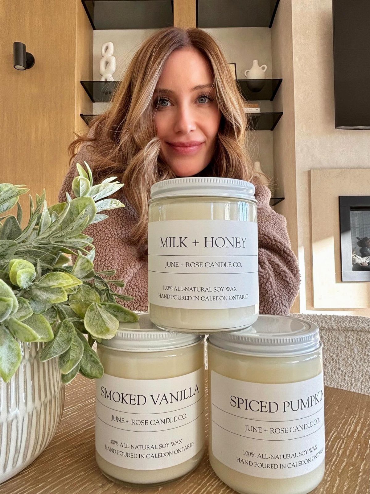 Three scented jar candles on a table with a smiling woman and plant.