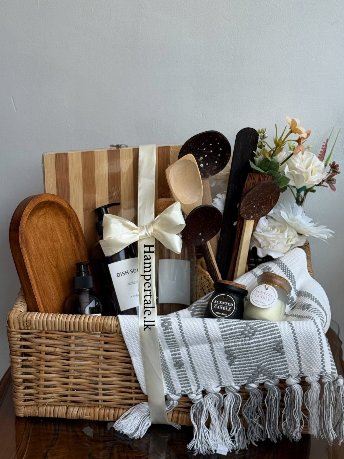 Woven gift basket with wooden kitchen utensils, cutting board, candles, and a textile.