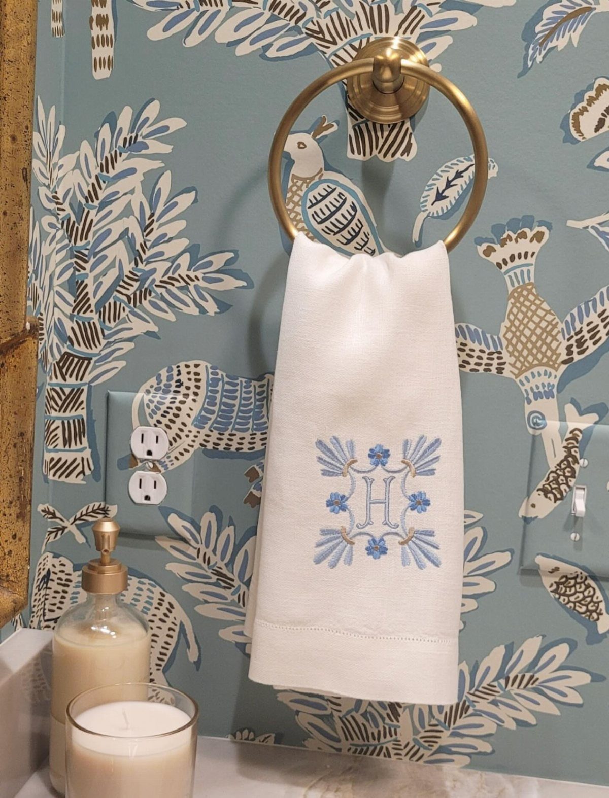 Monogrammed white hand towel hanging on a gold ring against patterned blue wallpaper.