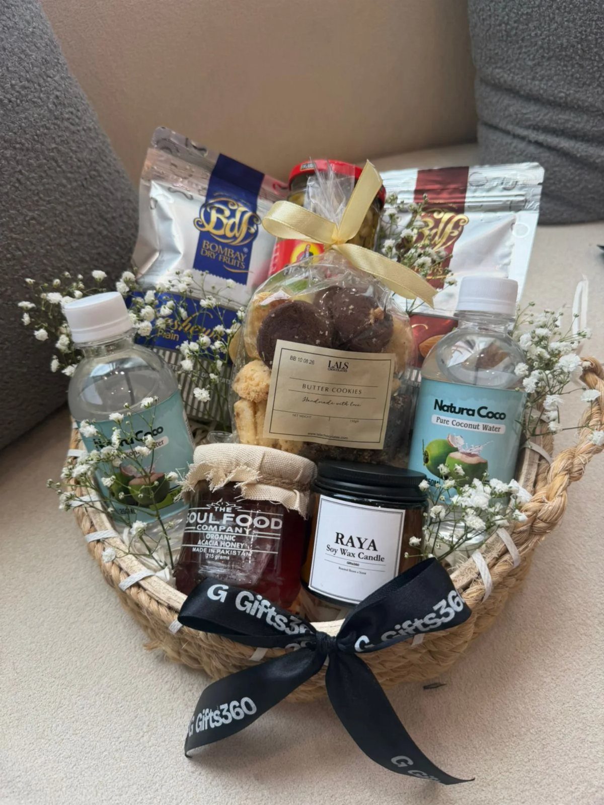 Wicker gift basket containing snacks, water, a candle, and jam with white flowers.