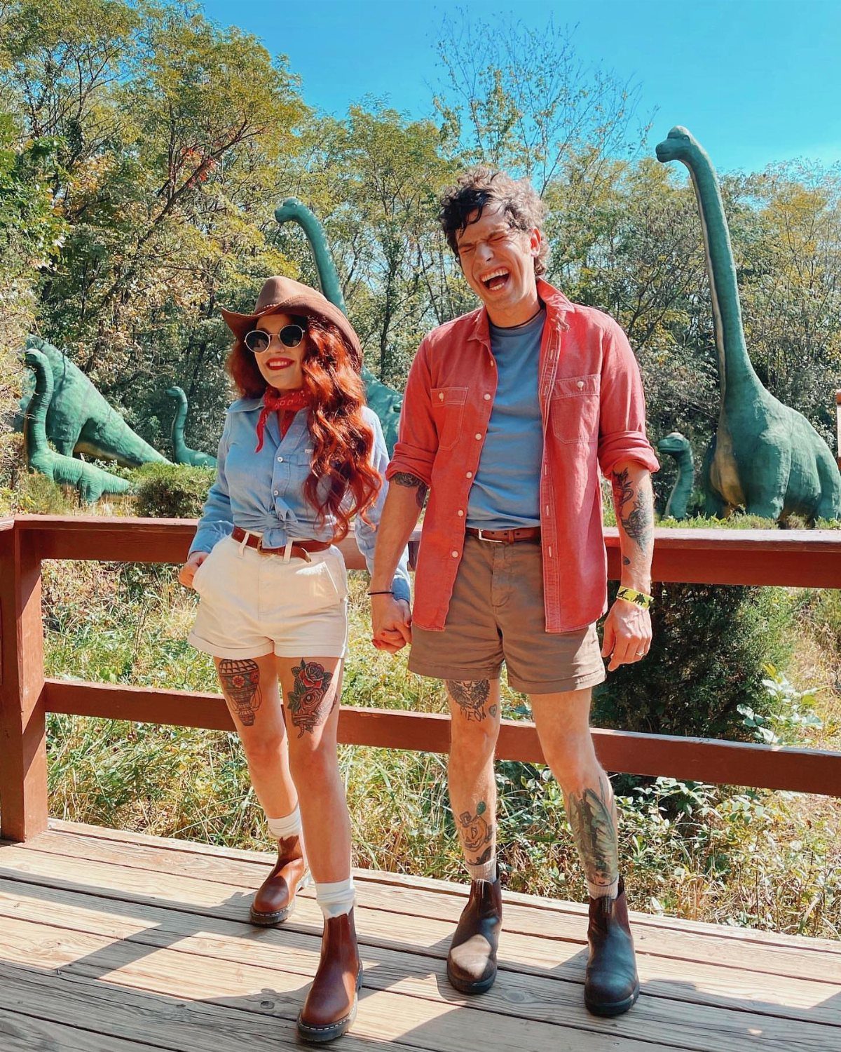 Couple dressed as Alan Grant and Ellie Sattler posing with dinosaur statues outdoors.