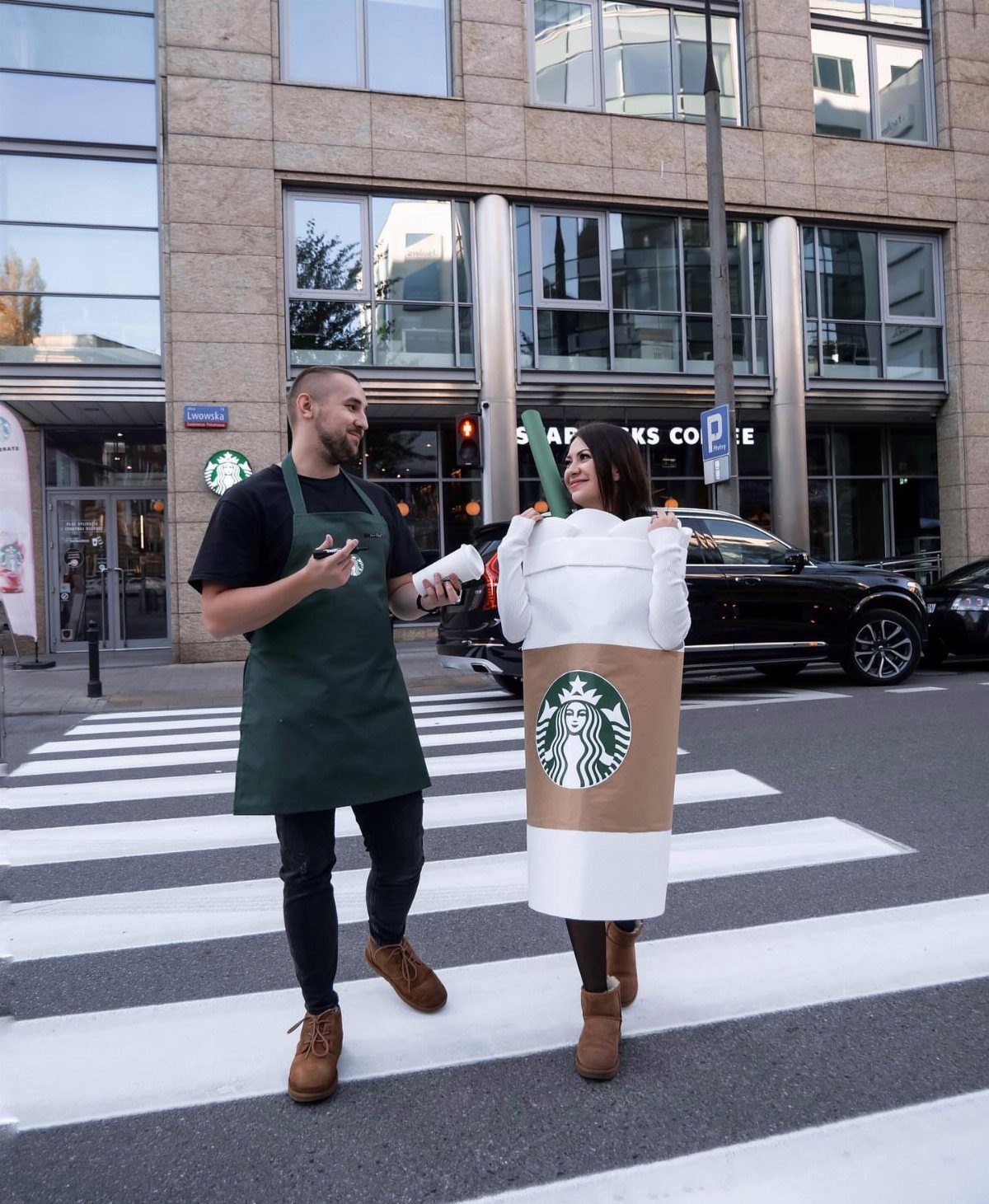 Man in green barista apron and woman in Starbucks cup costume crossing street.