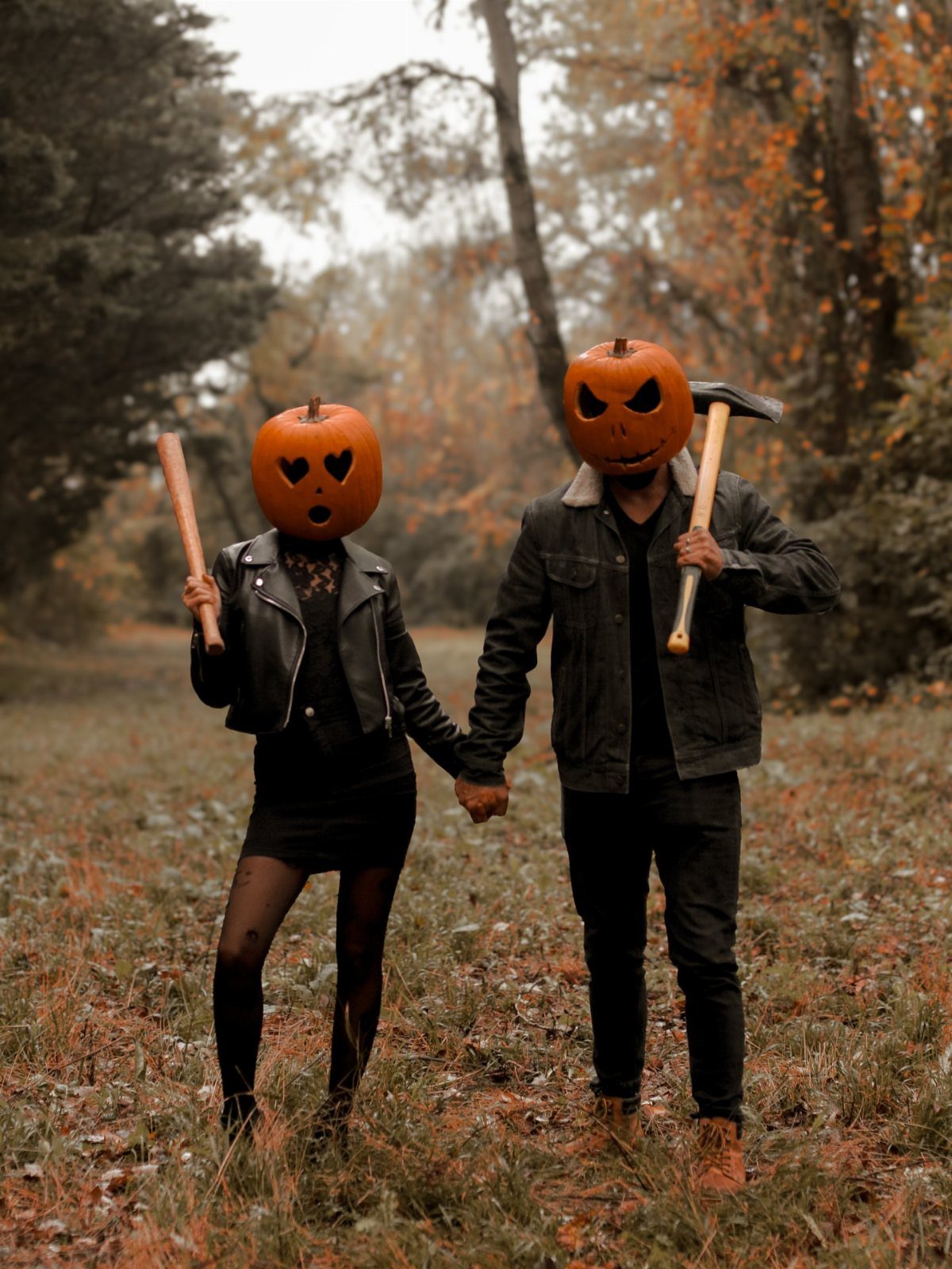 Couple with carved pumpkin heads holding hands in an autumn forest setting.