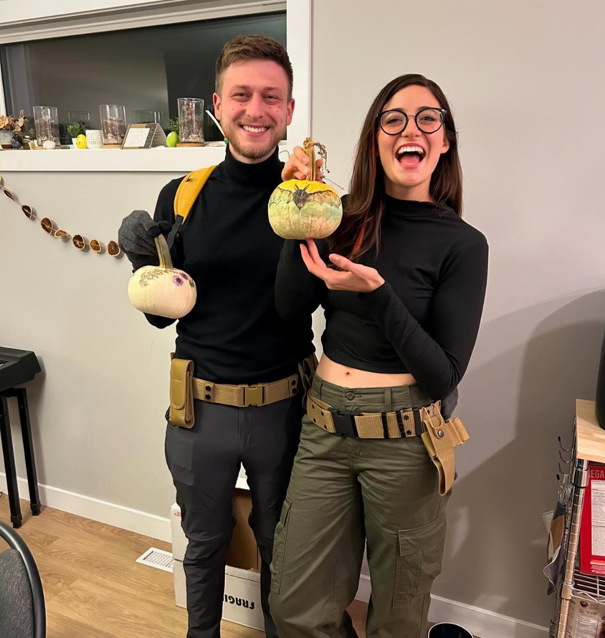 Man and woman in Kim Possible and Ron Stoppable costumes holding small pumpkins.