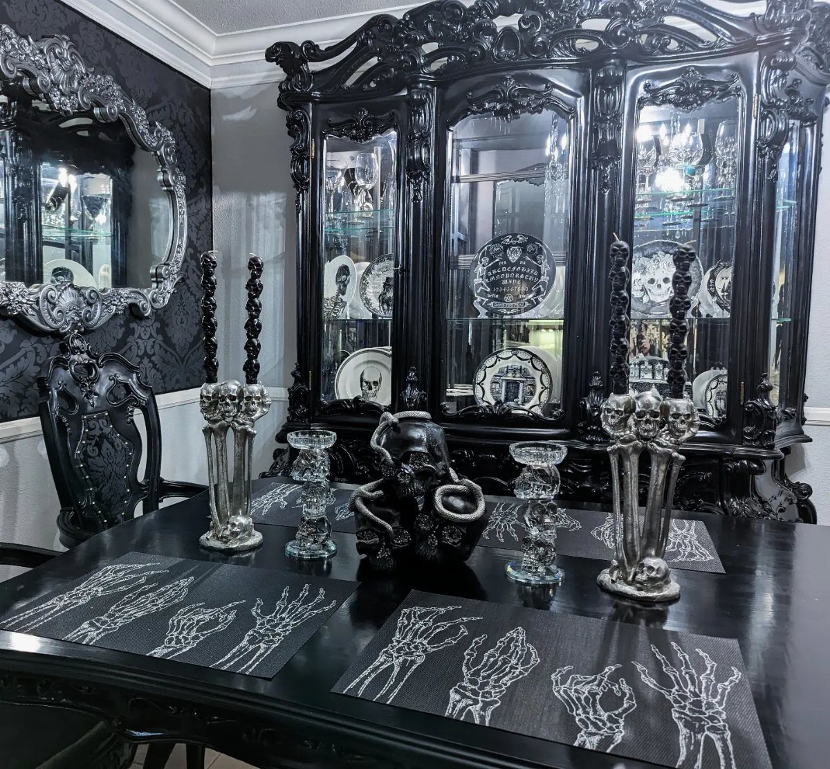 Gothic dining room with black ornate furniture, skull candle holders, and silver mirror.