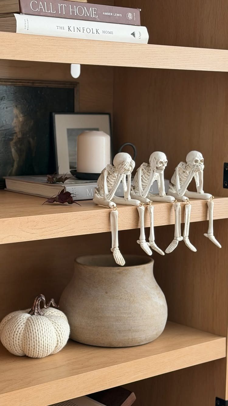 Three small skeletons sitting on a shelf above a white textured pumpkin and vase.