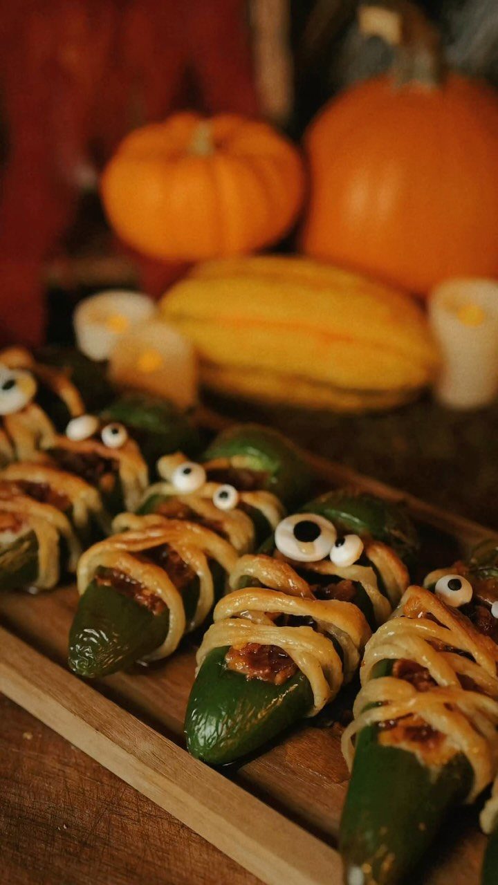 Jalapeno mummies wrapped in cheese with candy eyes on a wooden board.