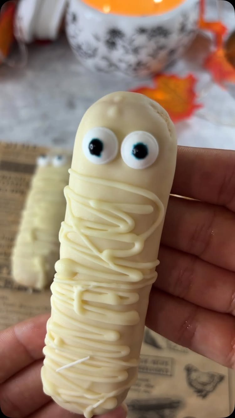 Mummy-shaped sausage snack with white cheese wraps and candy eyeballs held in hand.