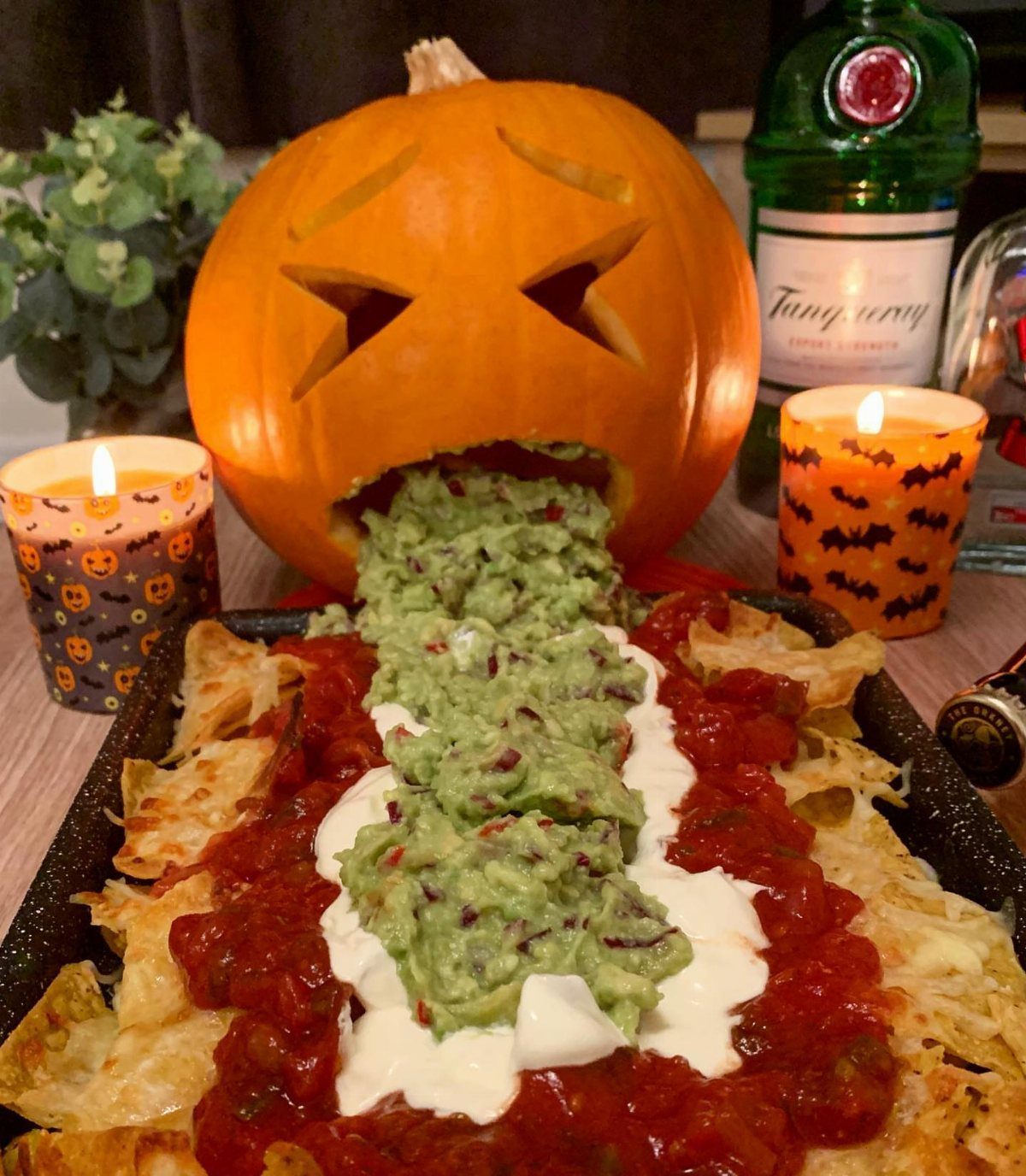 Carved pumpkin spilling guacamole onto a tray of nachos with salsa and cream.