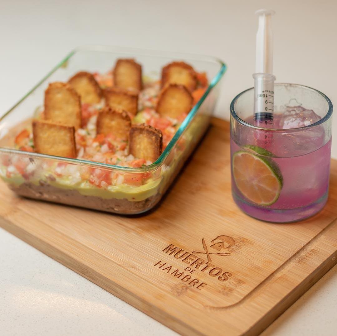 Layered taco dip with toast tombstones and a purple syringe cocktail on wood.