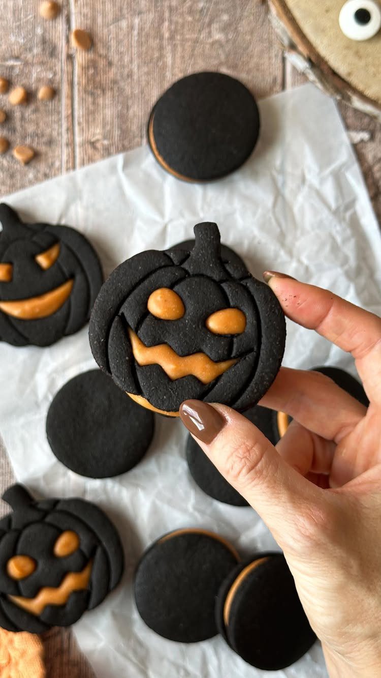 Black pumpkin-shaped cookies with orange frosting held by a hand on parchment paper.