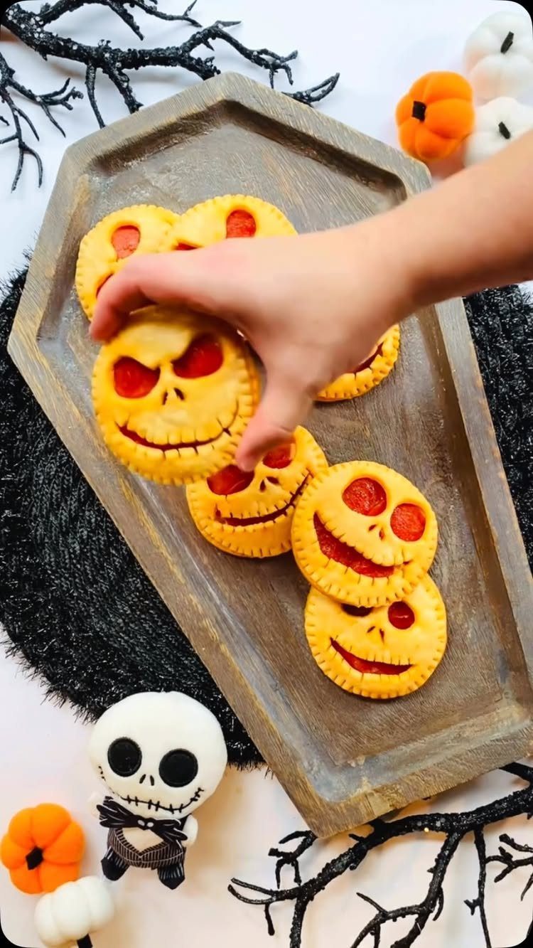 Jack-o'-lantern hand pies on a wooden tray with Halloween decorations and a hand.