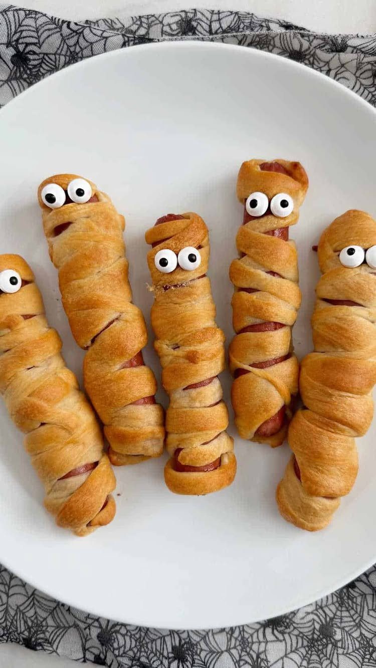 Mummy dogs wrapped in crescent dough with candy eyes on a white plate.