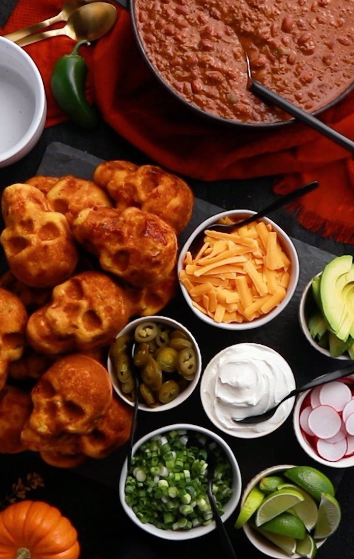 Skull-shaped cornbread muffins served with chili and various toppings on a dark surface.