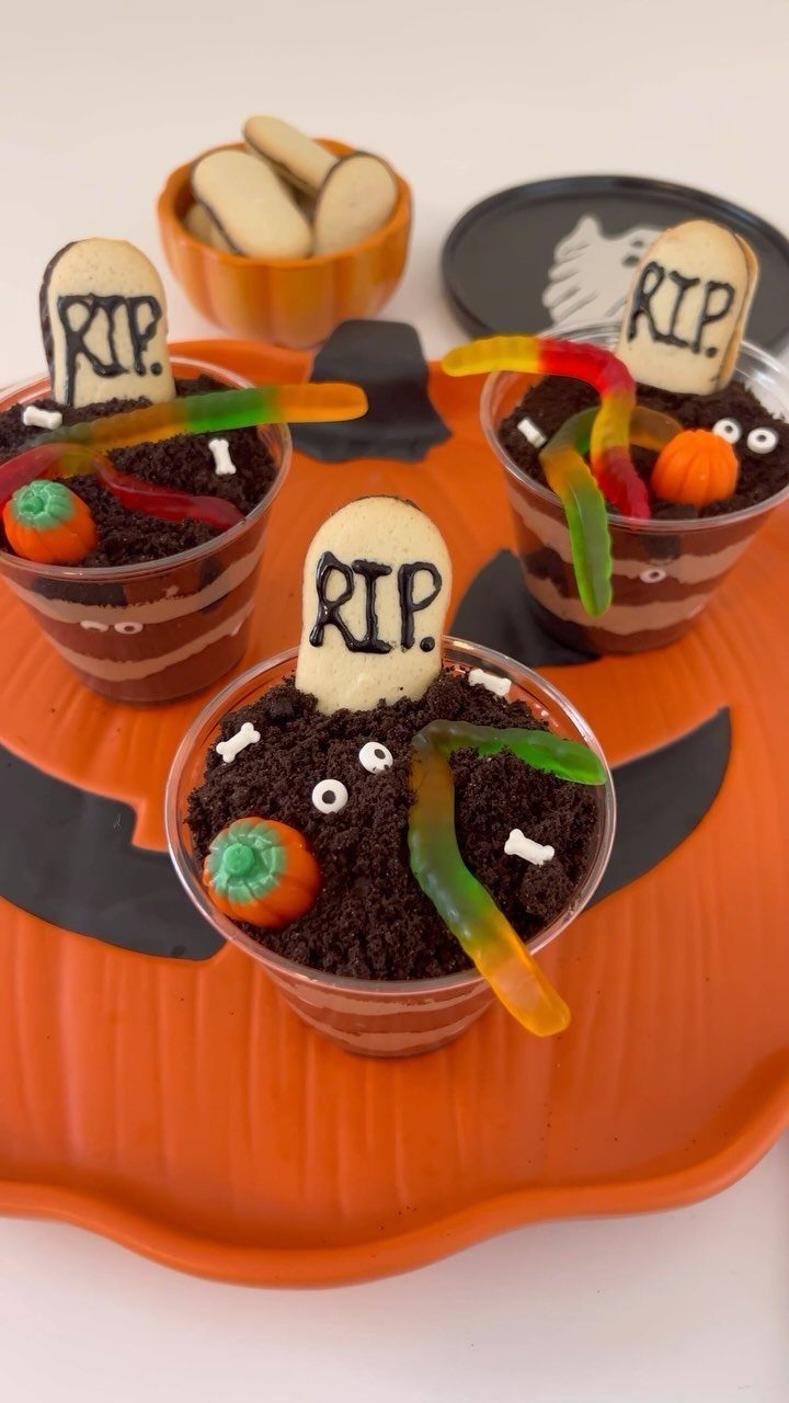 Graveyard pudding cups with cookie tombstones and gummy worms on a pumpkin platter.