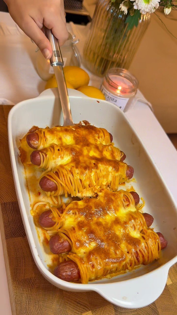 Hot dogs wrapped in spaghetti and melted cheese in a white baking dish.